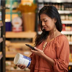 At a supermarket, a dark haired woman in orange blouse uses her mobile device to scan a QR code on a blue tin of food